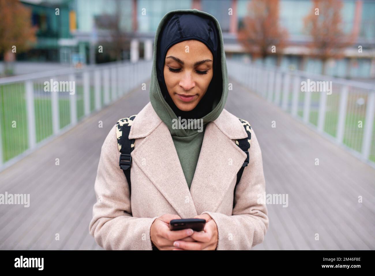 Portrait of young muslim woman in coat, standing outdoor in city, scrolling smartphone. Stock Photo