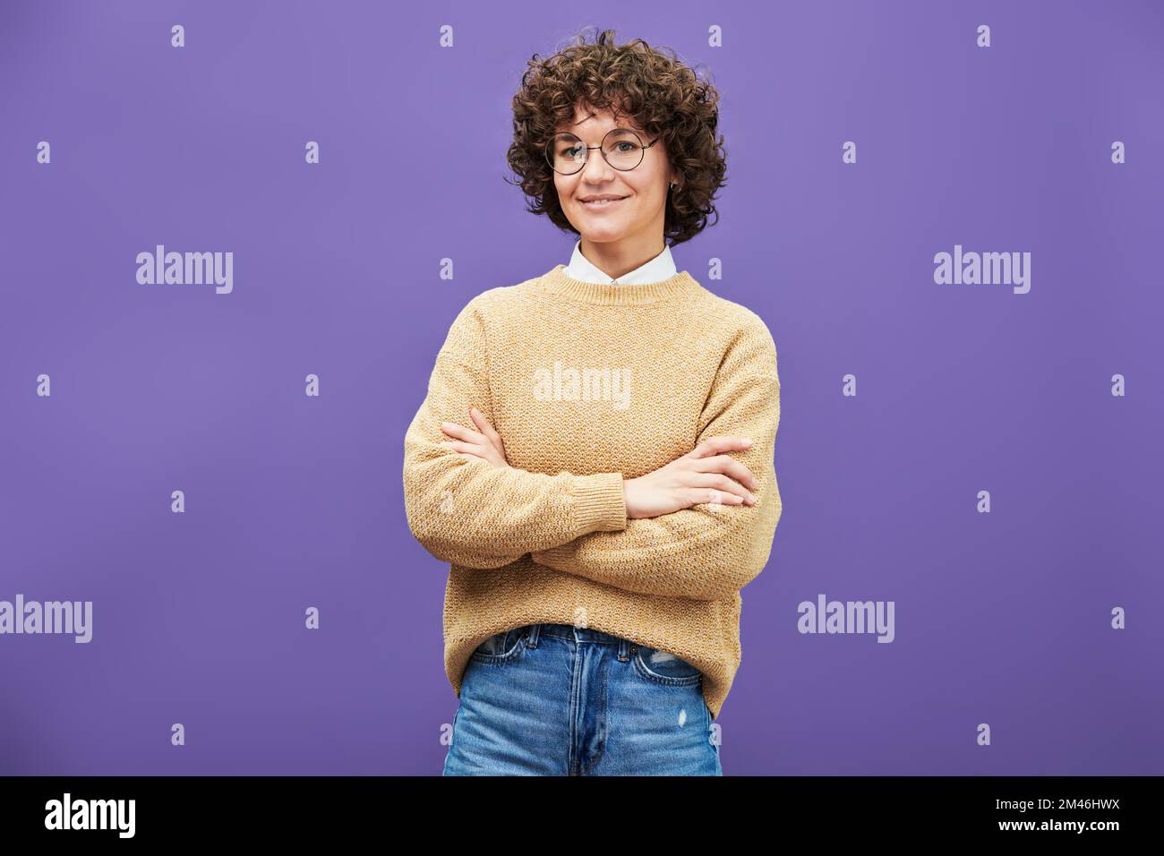 Young smiling brunette woman in eyeglasses, blue jeans and beige knitted pullover crossing arms by chest and looking at camera in isolation Stock Photo