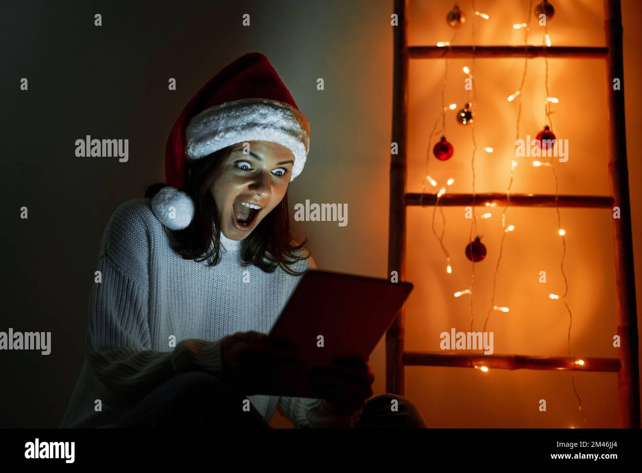 Happy woman in Santa hat holding digital tablet in her hands looking at screen and screaming with happiness and surprise. Stock Photo