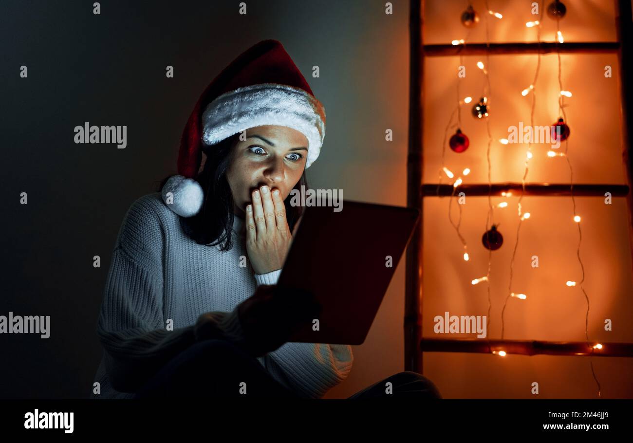 Woman wearing Santa hat looking at the screen of digital tablet confused covering her mouth with hand. Stock Photo
