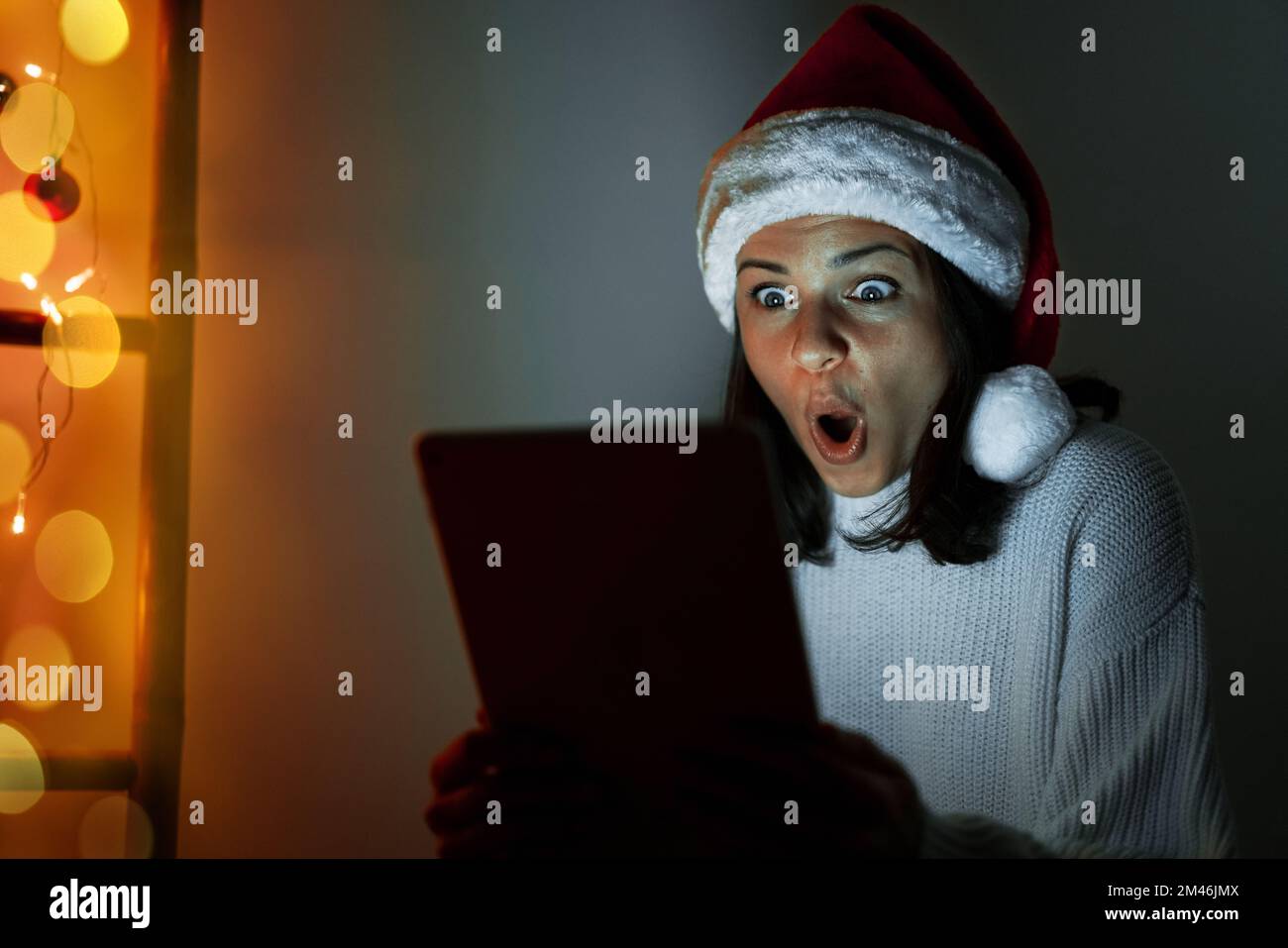 Woman in Santa hat holding digital tablet in her hands and looking at screen in surprise with wide eyes. Stock Photo