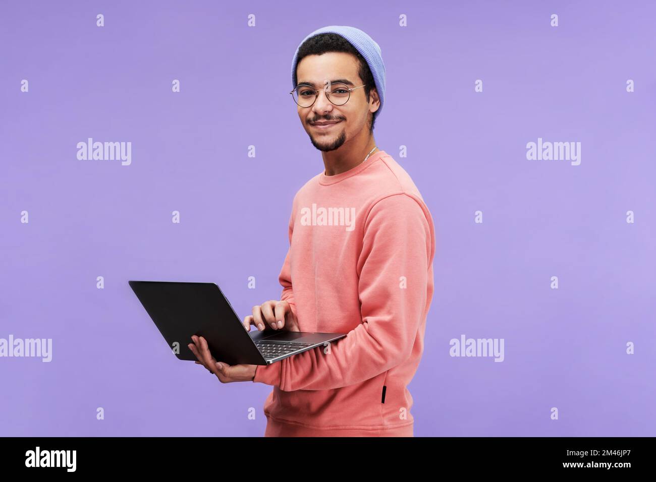Young successful male freelancer or programmer in pink pullover, beanie hat and eyeglasses holding laptop and looking at camera Stock Photo