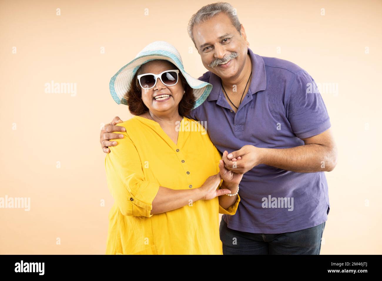 Portrait of Happy indian senior couple wearing hat and sunglasses and casual cloths standing together over beige background. Retired old husband and w Stock Photo