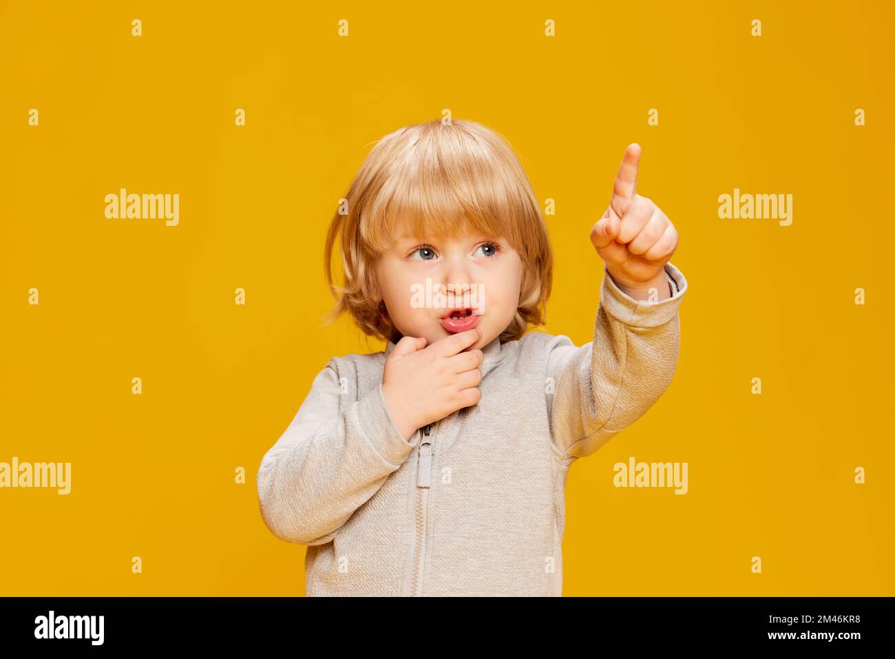 Portrait of cute little boy, child posing, pointing somewhere over yellow studio background. Curious kid. Concept of childhood, emotions Stock Photo