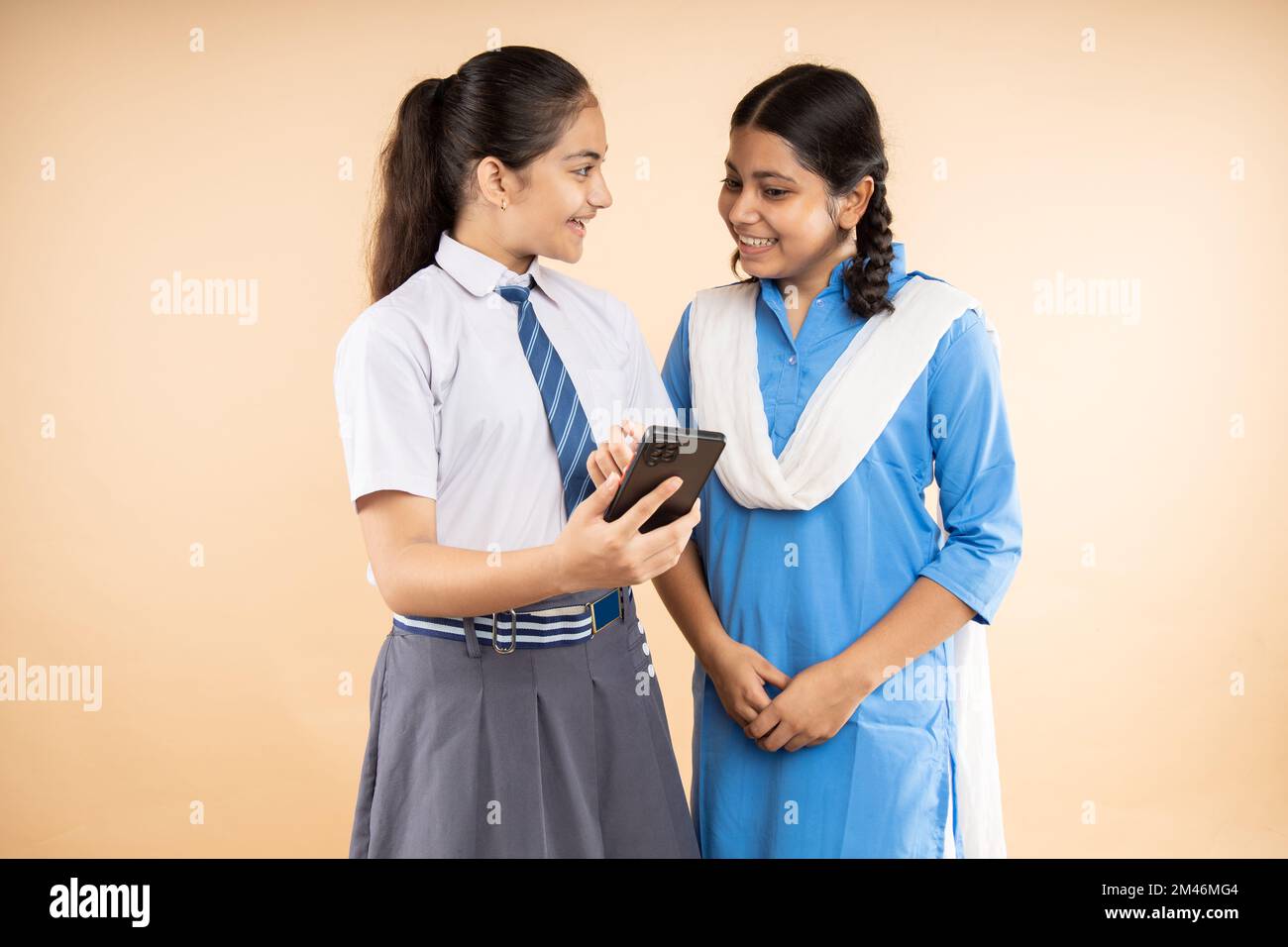 Happy Rural and Modern Indian student schoolgirls wearing school uniform using smart phone together isolated over beige background, Closeup, Studio sh Stock Photo