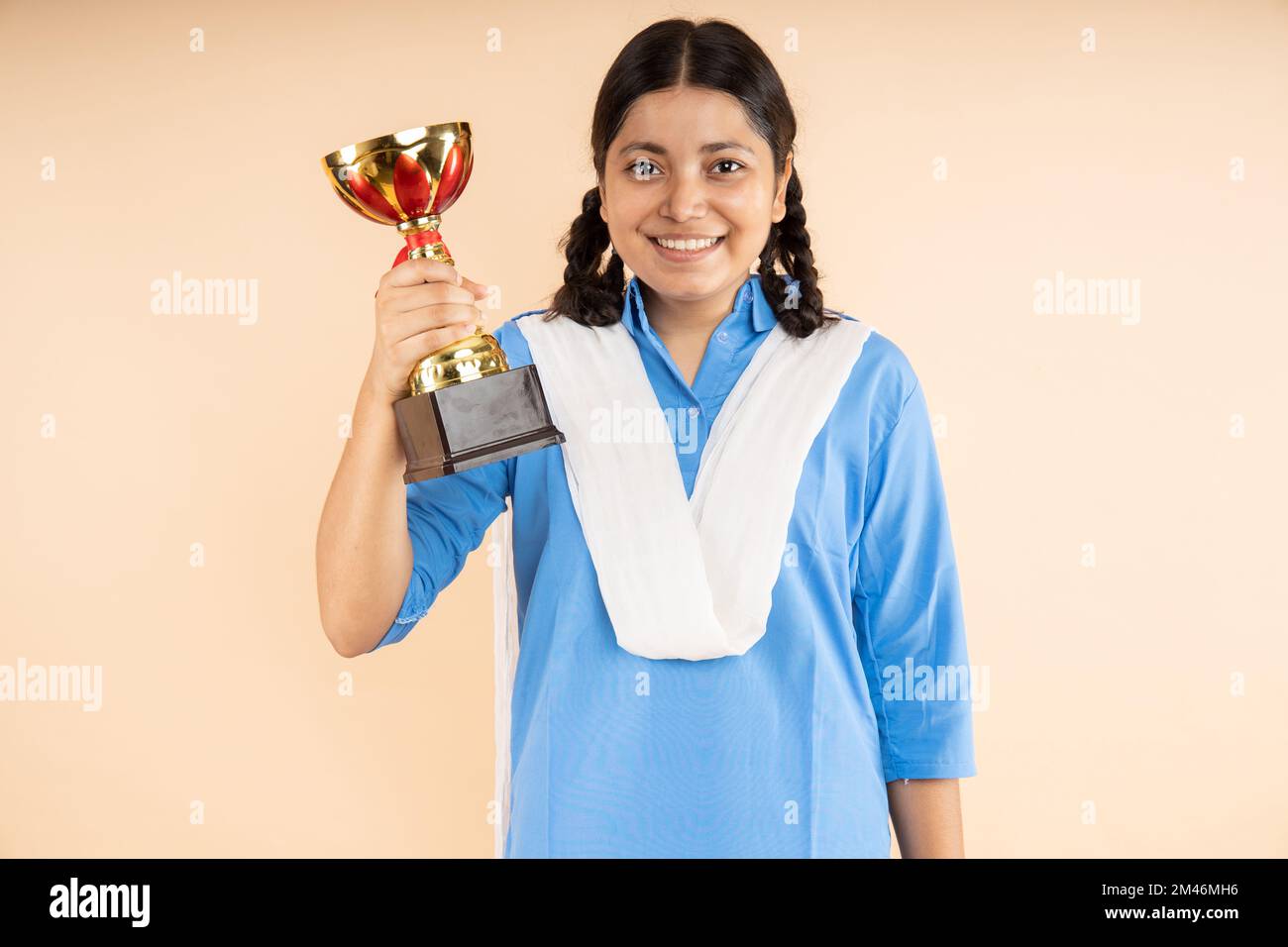 Happy Rural Indian student schoolgirl wearing blue government school uniform hold victory trophy in hand isolated over beige background, winner,Studio Stock Photo