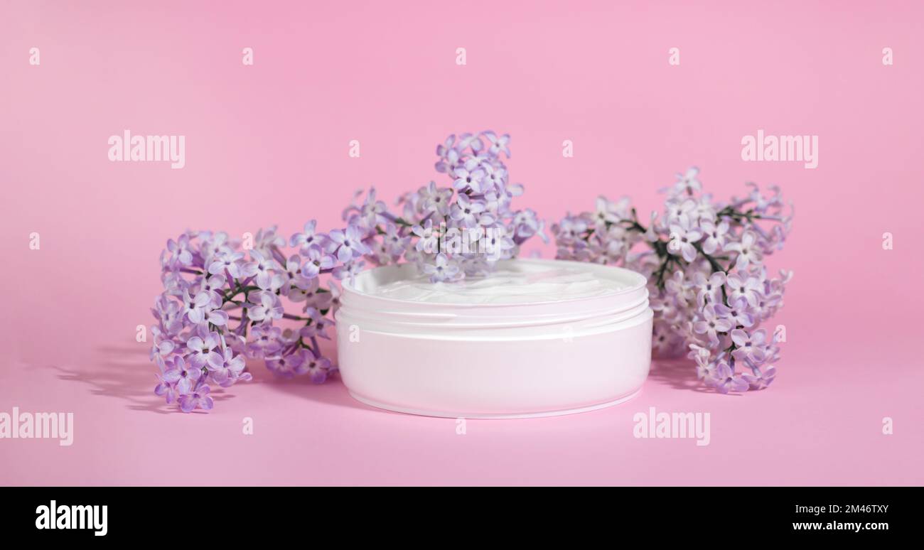 White jar of moisturizing face cream with pink cap isolated on pink background with lilac flowers. eco cosmetology or skin care concept. Stock Photo