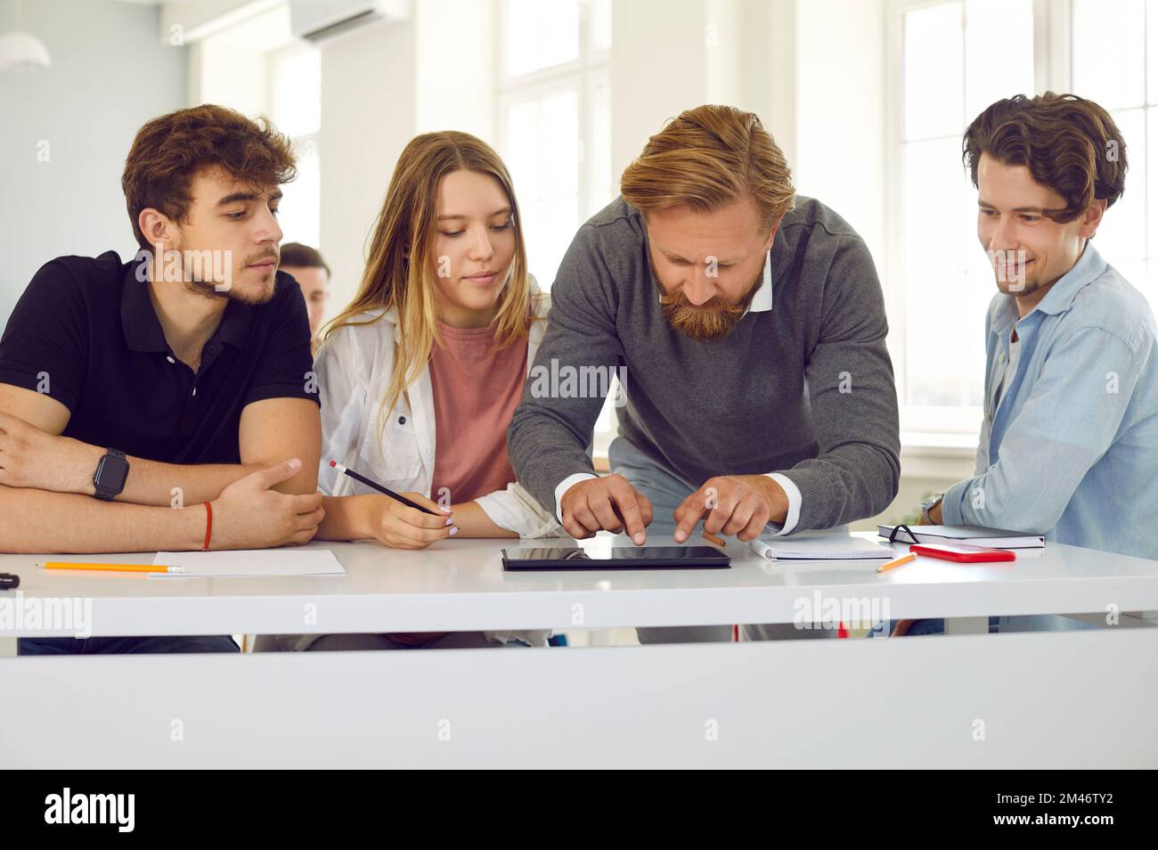 Male teacher in class helps group of highschool students working on digital tablet. Stock Photo