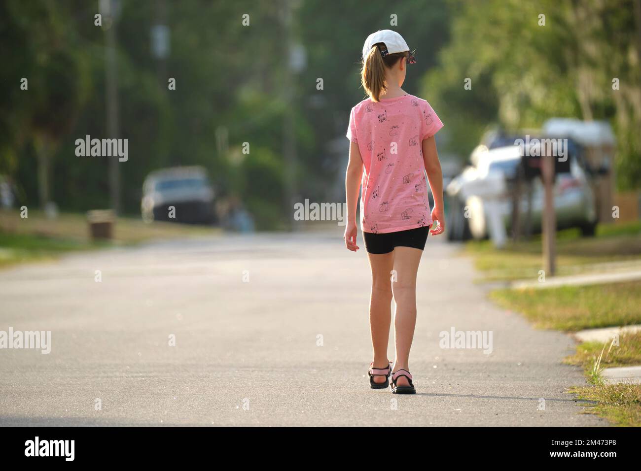Rear view of confident young child girl walking along the sunny alley ...
