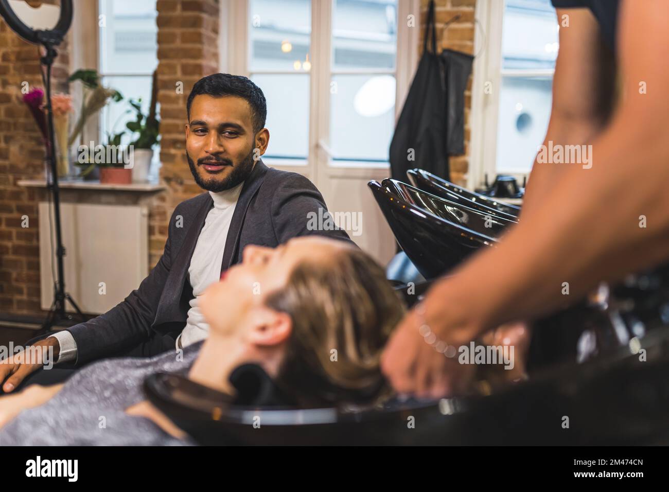 Smiling husband looking at his wife having her hair washed by expert hair stylist at their favorite hairdresser place. High quality photo Stock Photo