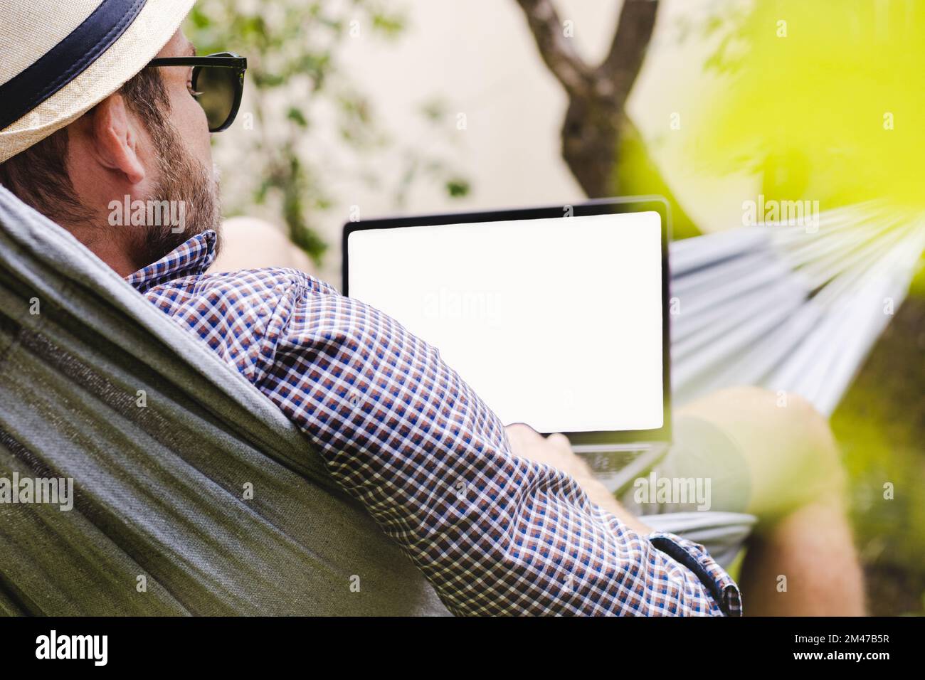 Businessman in hat and sunglasses lying down on hammock and using his laptop. Stock Photo