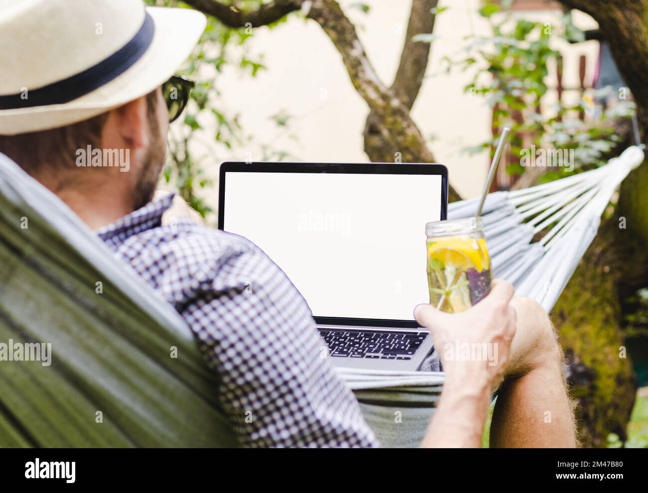 Laptop with blank screen and rear view man lying down in a hammock with a glass of water in his hand. Stock Photo
