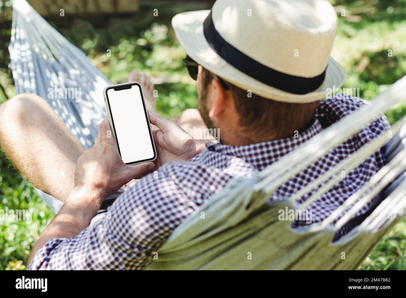 Man in hat using his smartphone while lying in hammock and relaxing. Stock Photo