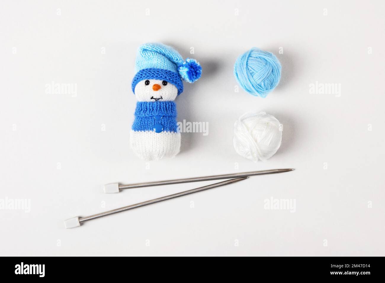 Knitted snowman in a blue hat and sweater with two balls of thread and knitting needles on a gray background. Knitted Christmas decorations Stock Photo