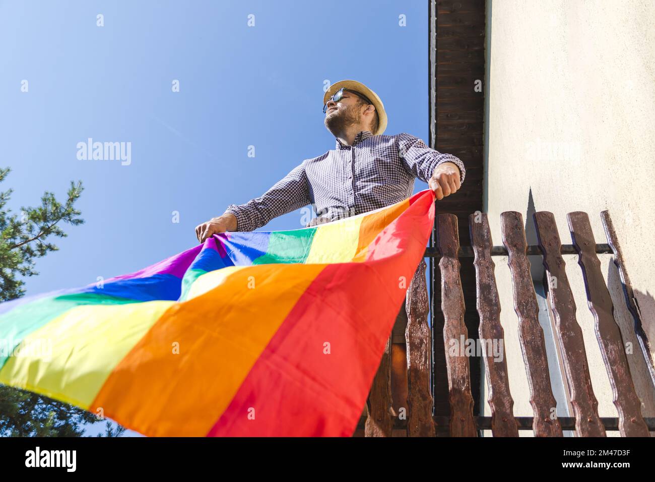 Handsome bearded man in hat standing on the balcony and holding rainbow flag, pride month, equality, equity, diversity. Stock Photo