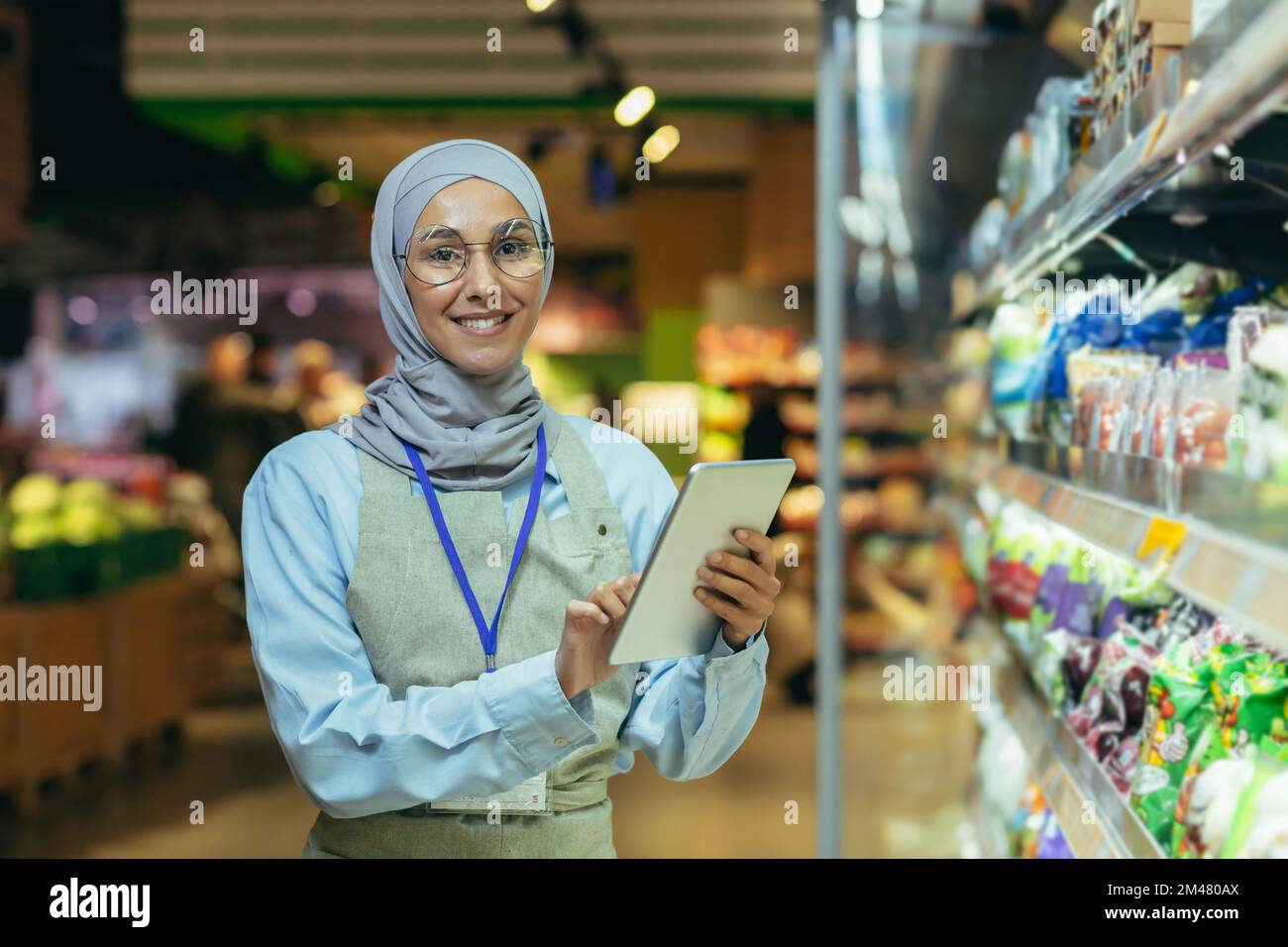 Portrait of happy and smiling seller woman in hijab, muslim woman with tablet smiling and looking at camera, saleswoman near field with vegetables and salad products chooses and sifts term. Stock Photo