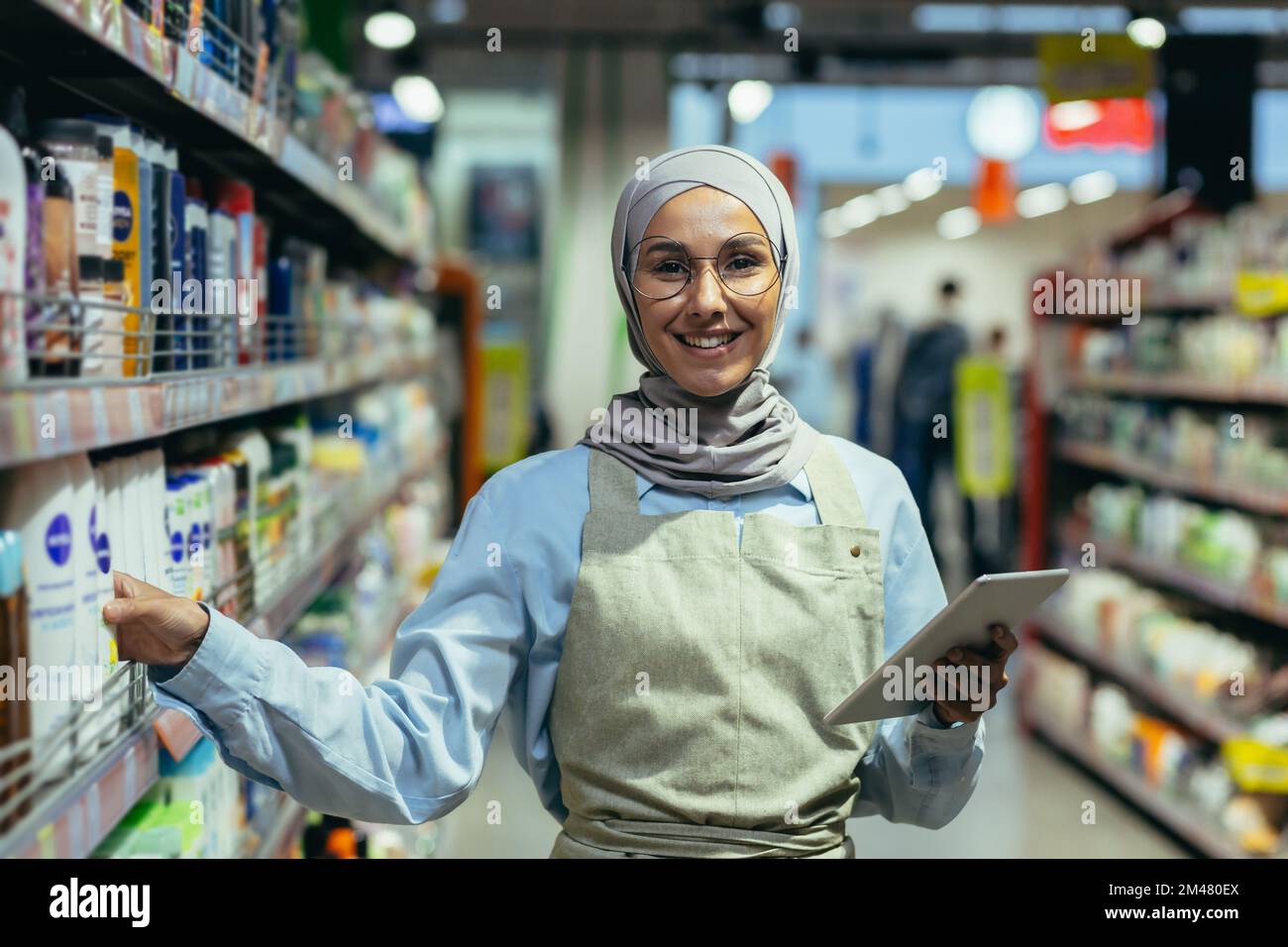 Portrait of a female saleswoman in a hijab, a salesperson in a household chemicals department is smiling and looking at the camera, holding a laptop tablet computer in her hands. Stock Photo
