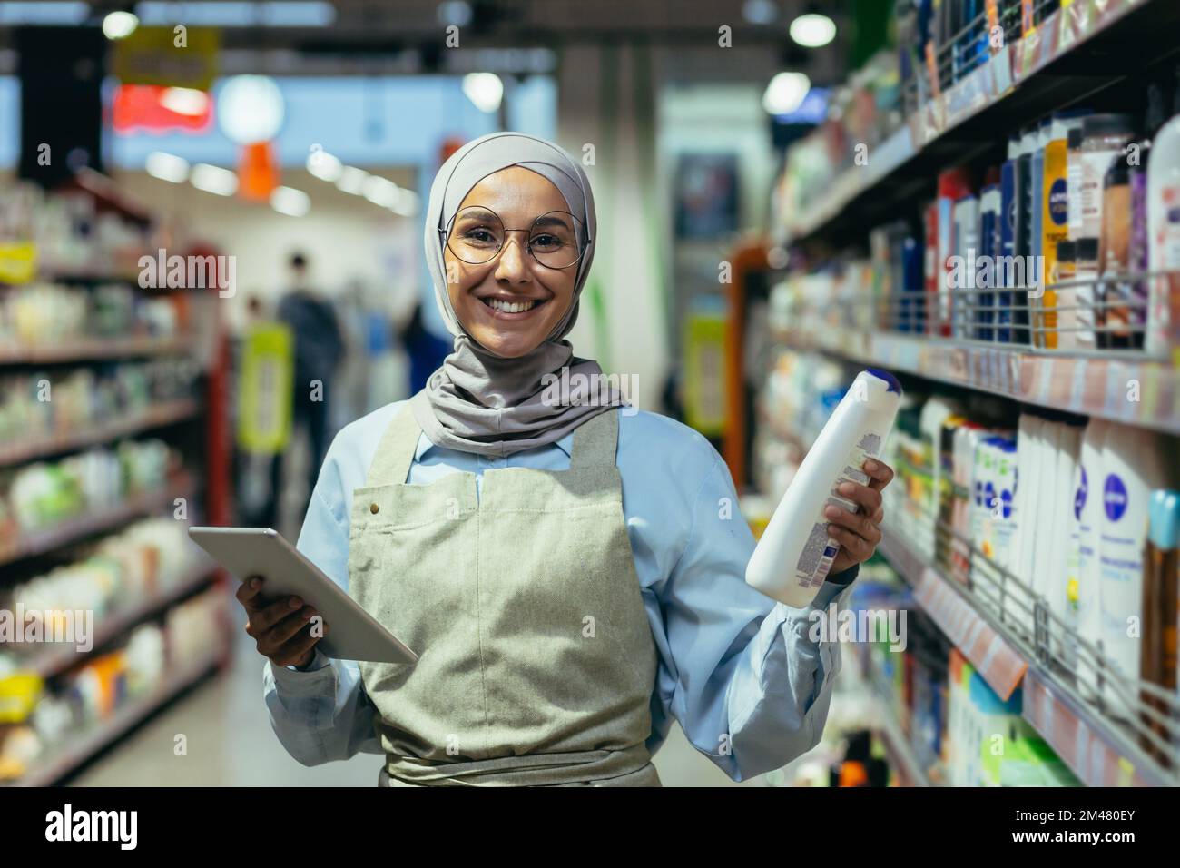 Portrait of a female saleswoman in a hijab, a salesperson in a household chemicals department is smiling and looking at the camera, holding a laptop tablet computer in her hands. Stock Photo