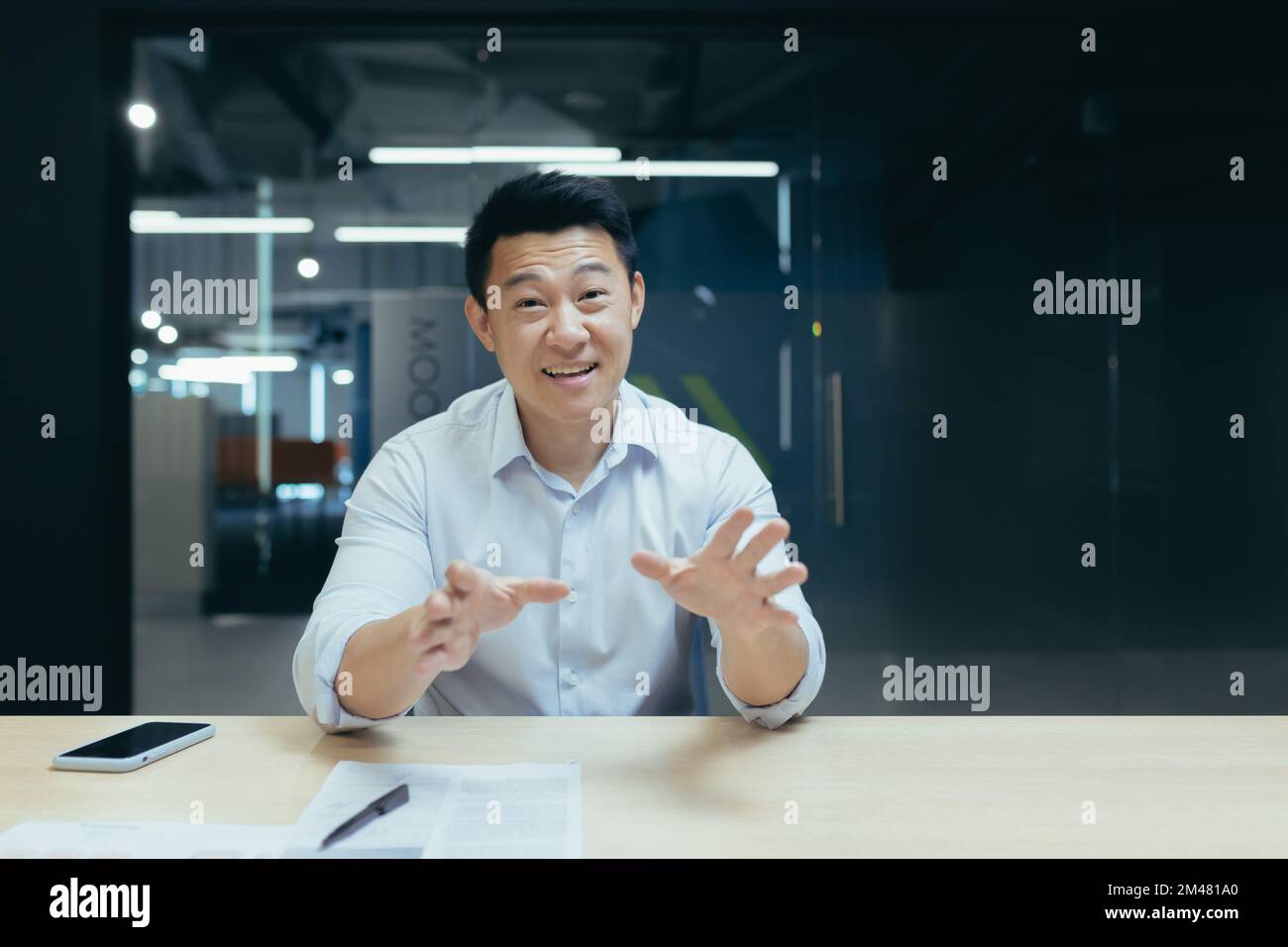 Smiling young Asian man sitting in the office at the desk and talking to the camera. Conducts online meetings, consults, business training. Stock Photo