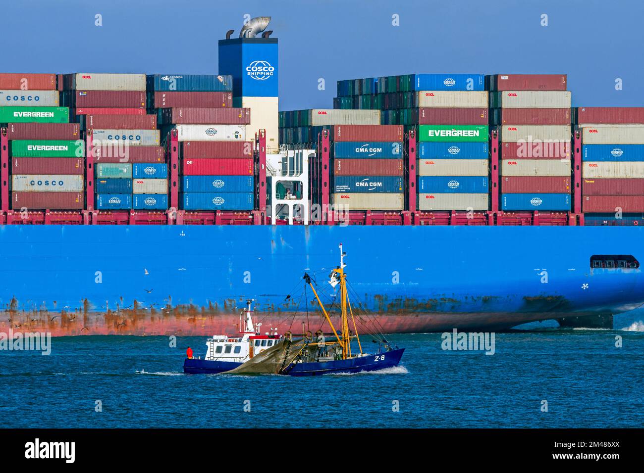 Little fishing boat passing huge Chinese COSCO Shipping container ship loaded with containers on the North Sea, sailing under flag of Hong Kong, China Stock Photo