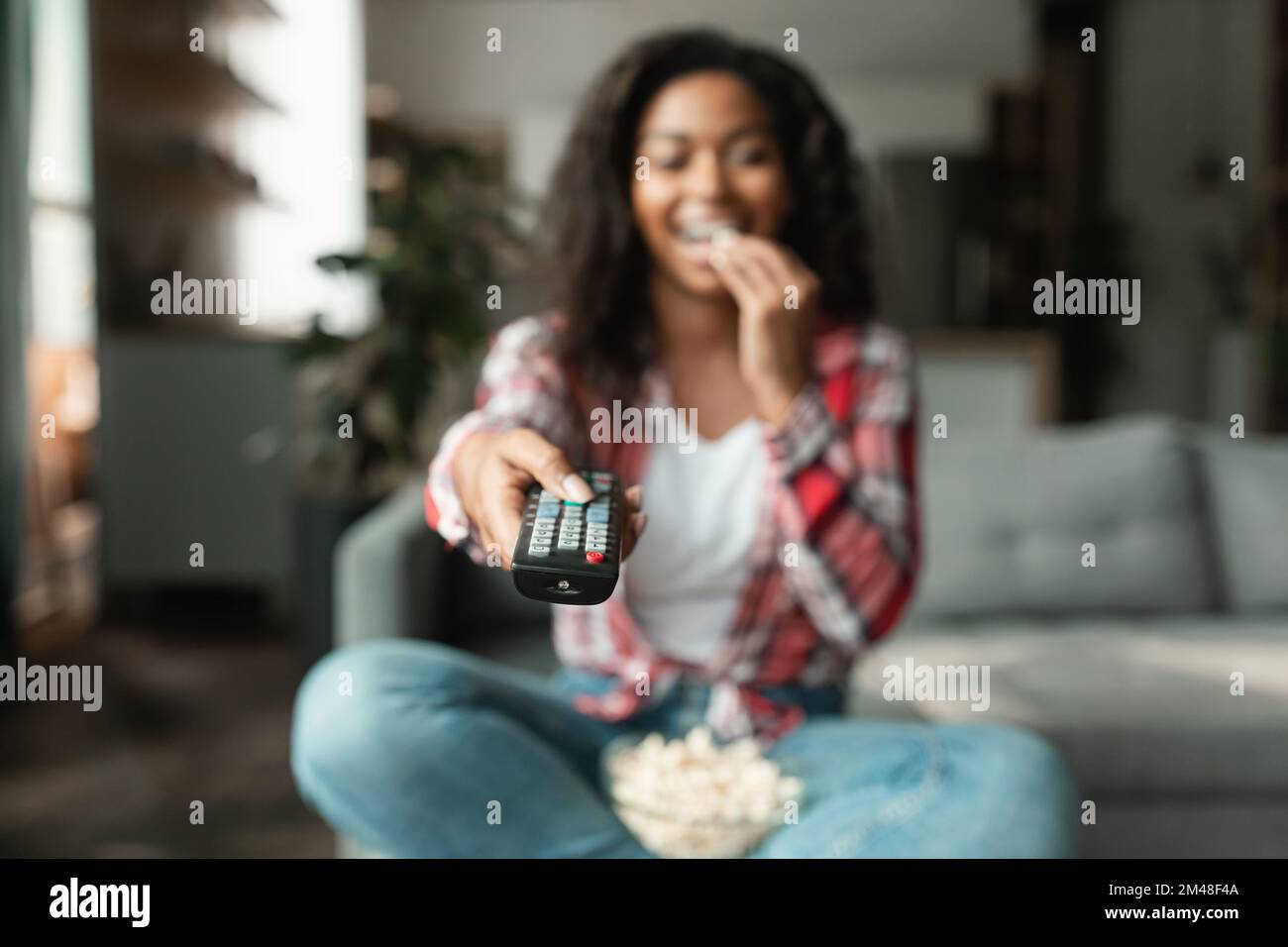 Happy millennial black lady with remote control change channel on tv, watch movie, eat popcorn, enjoy free time Stock Photo