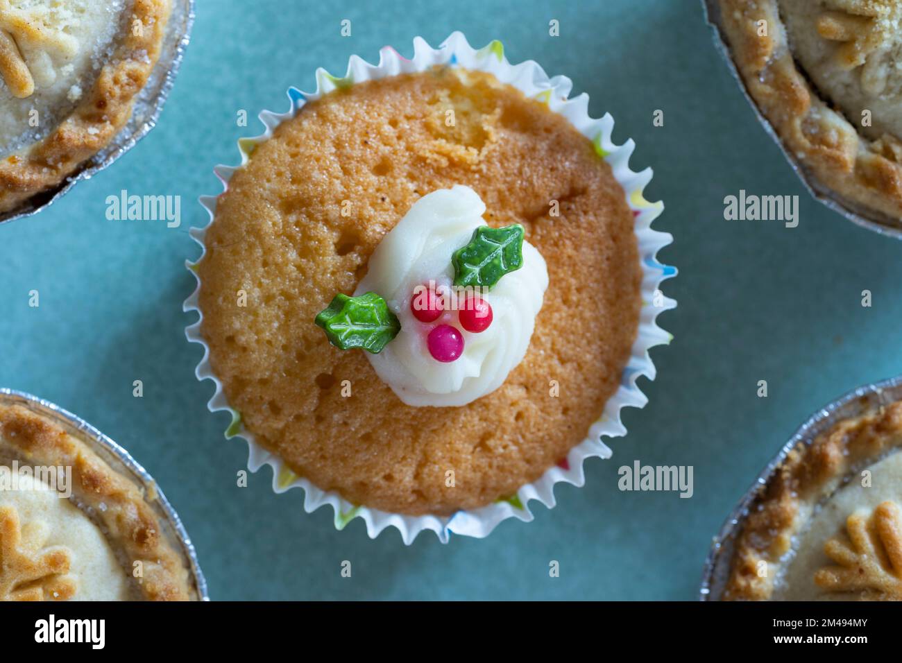 Home made cup cake with white icing and edible green holly leaves and red berries on a blue plate at Christmas time, UK Stock Photo