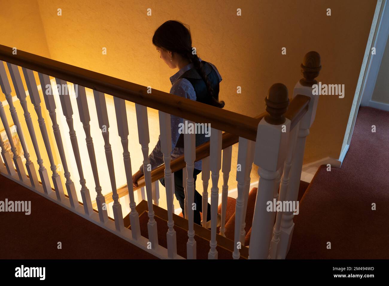 A young woman walking downstairs in a UK house. Concept: feeling alone, feeling fearful, feeling trepidation, feeling alone, walking into the light Stock Photo