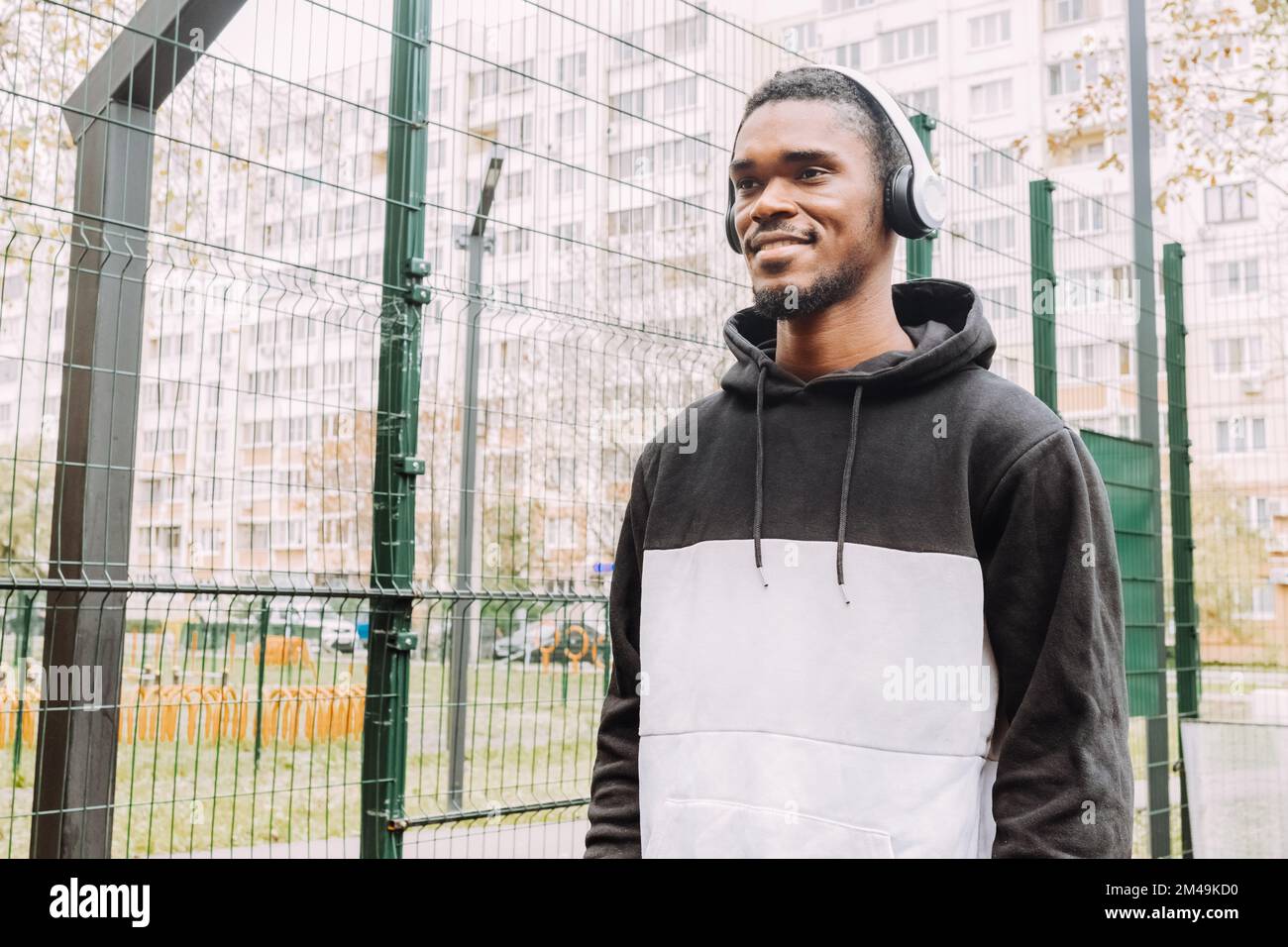 Cheerful handsome black guy in wireless headphones on sport court. Smiling young man in two-tone hoodie in earphones. Bottom view. Stock Photo