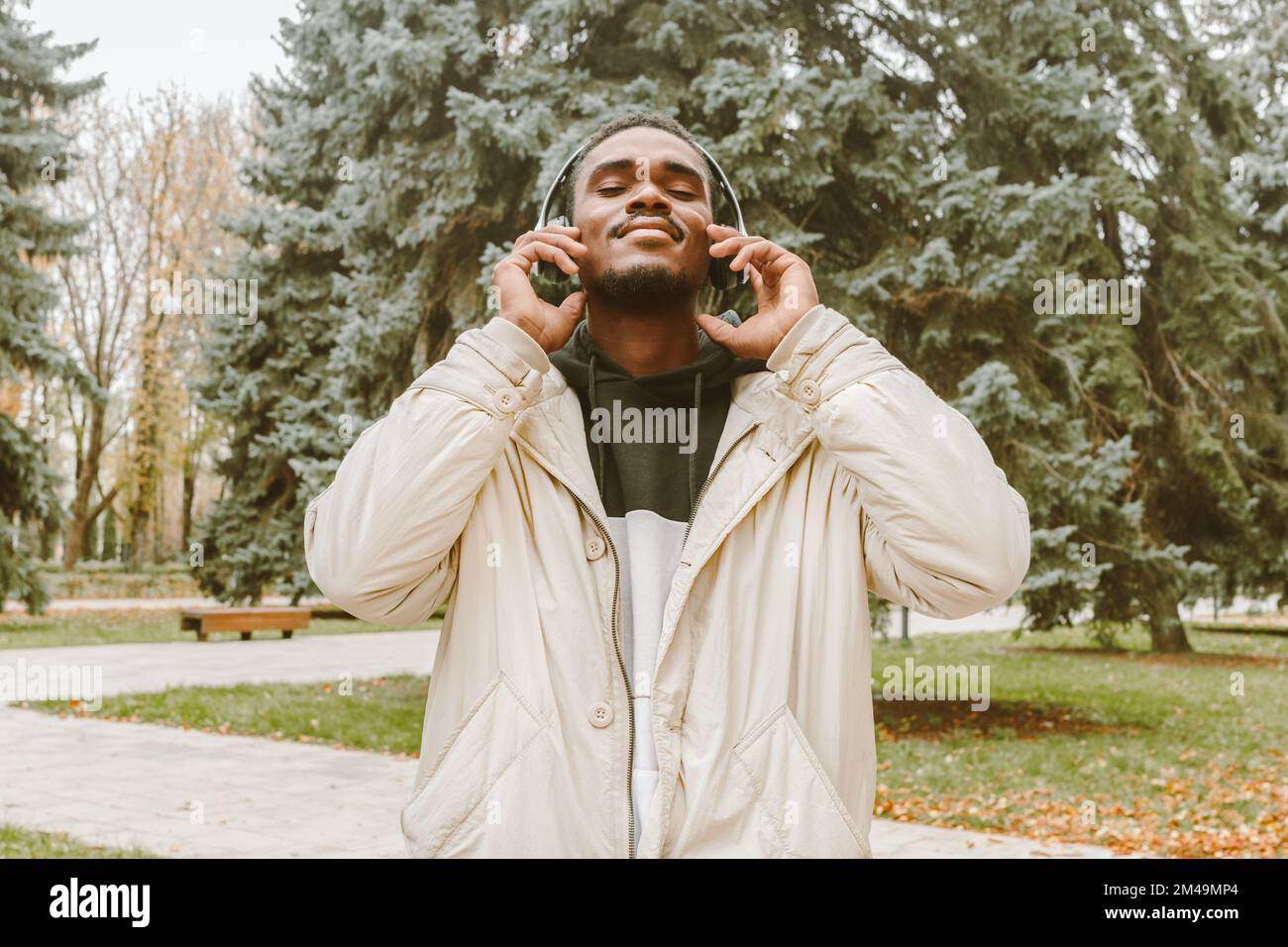 Portrait of handsome black man in wireless headphones over park backdrop. Smiling African guy with closed eyes in warm clothes and in white earphones Stock Photo
