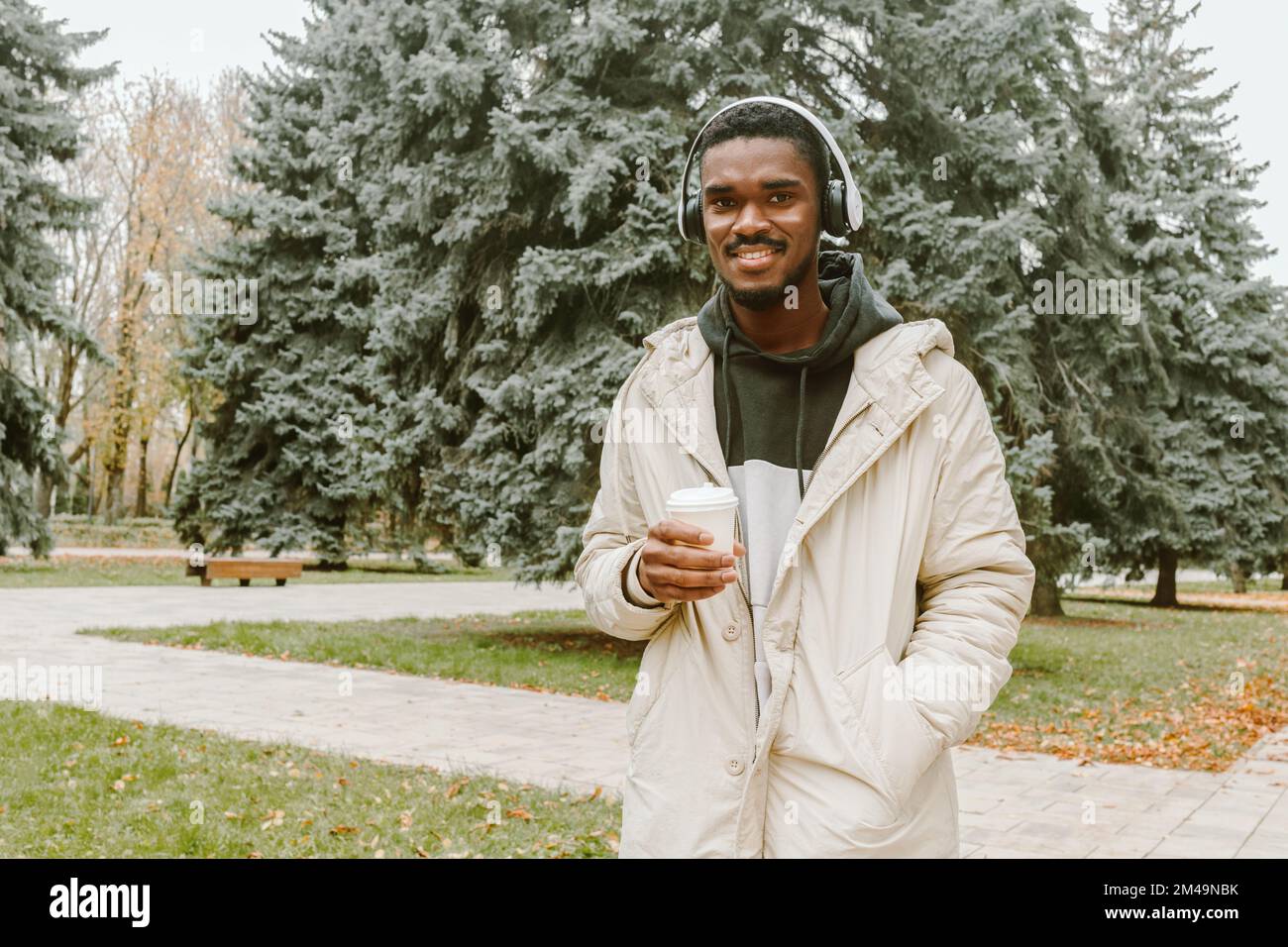 Young black man walking in park with cup of coffee on autumn day. Smiling guy wearing casual warm clothes in wireless headphones Stock Photo