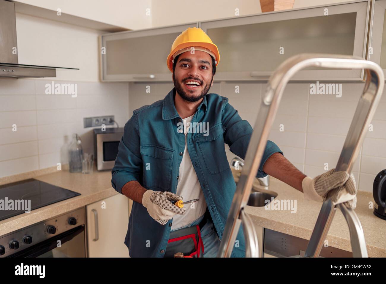 Smiling handyman is standing on ladder in kitchen and holding a screwdriver Stock Photo