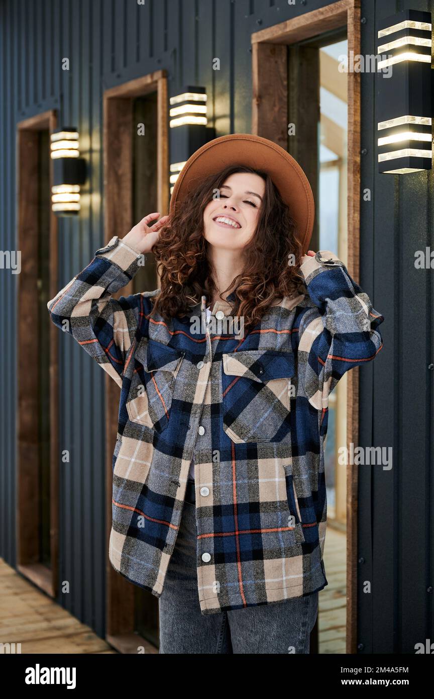Curly young woman resting on terrace of modern barn house in the mountains. Happy female tourist enjoying winter holidays in new cottage. Stock Photo
