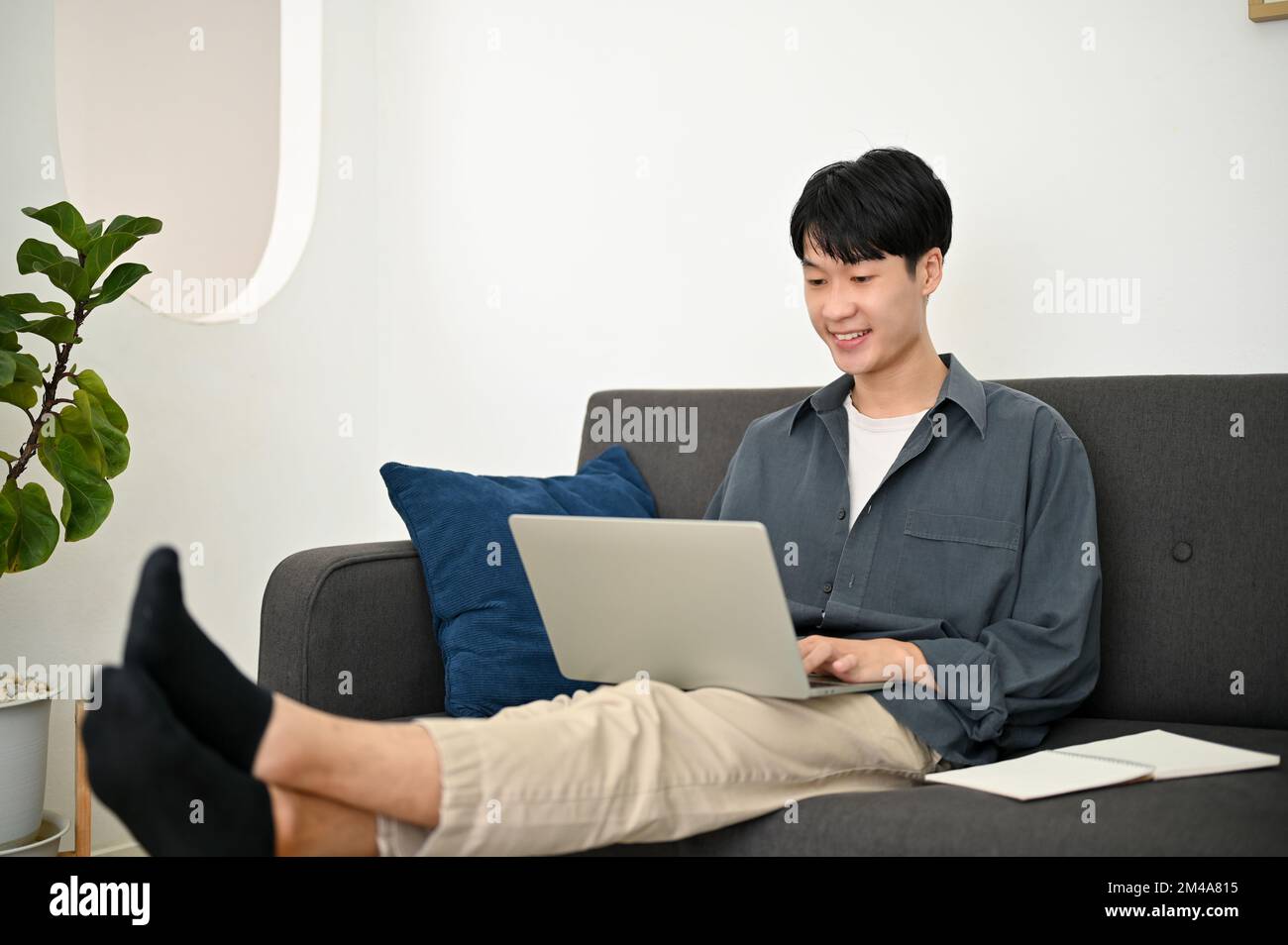 Relaxed and chilling young Asian man remote working from home, using laptop while sitting on sofa in the living room. Stock Photo