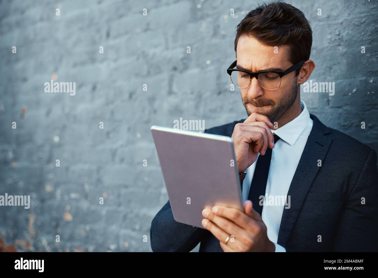 Giving it some thought. a handsome young businessman using his tablet while standing against a grey facebrick wall. Stock Photo
