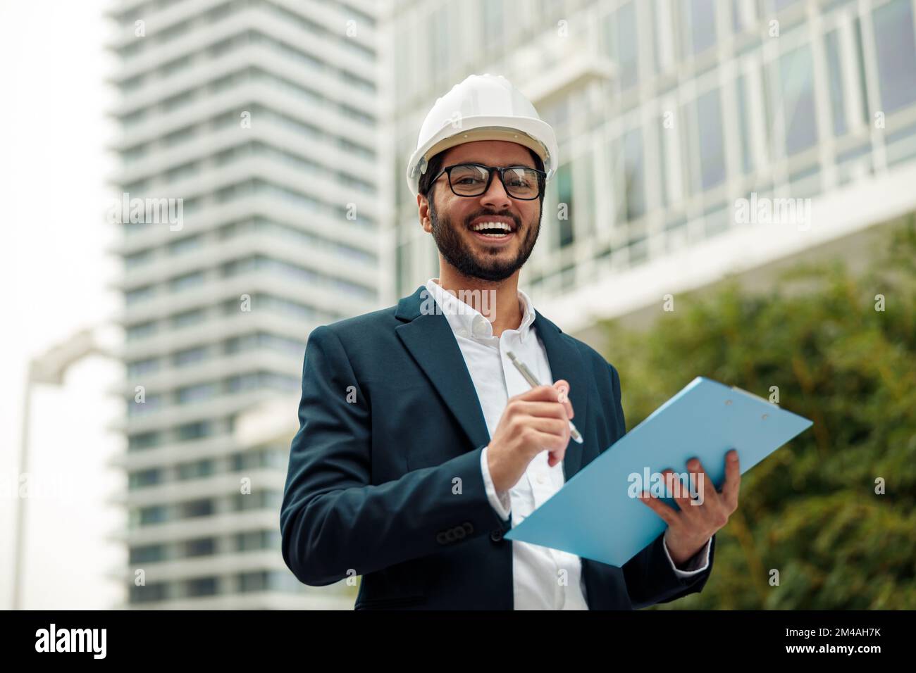 Indian construction architect in suit and hardhat making notes against modern building Stock Photo