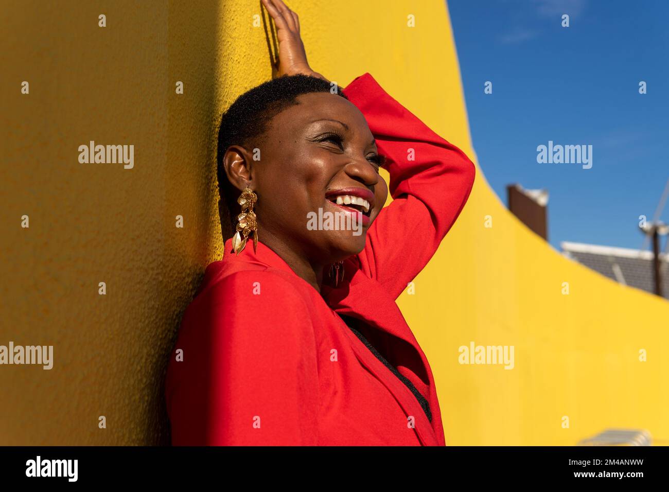 Side view of smiling young stylish adult African American female with short dark hair in stylish suit and earrings looking away standing near yellow w Stock Photo