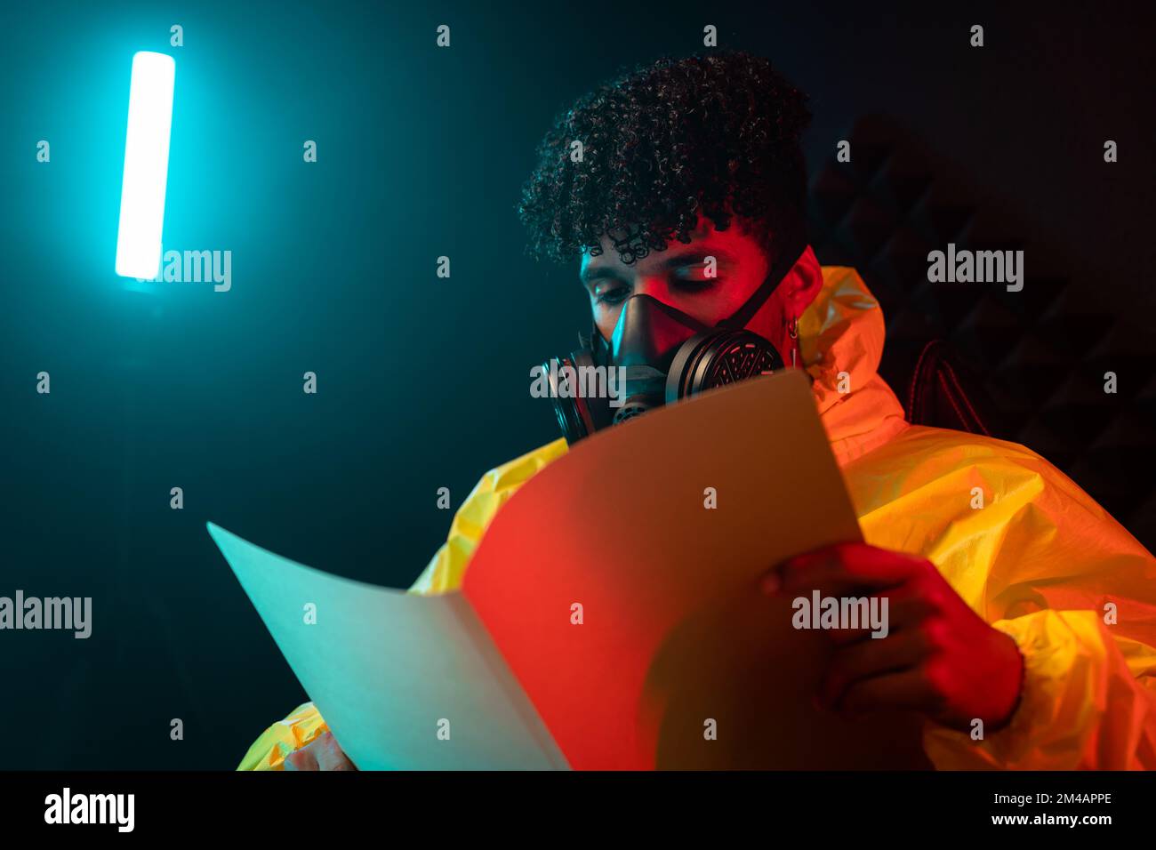 Young Hispanic man in yellow hazmat suit and gas mask reading papers in