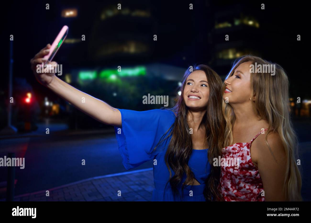 Happy young brunette in stylish blue dress smiling and taking selfie with mature mom while standing on city street after party at night Stock Photo