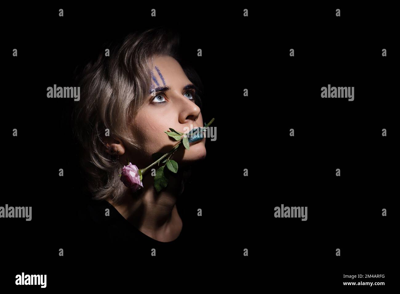 dark dramatic female portrait with trendy teal color makeup hold a rose flower in her mouth. blond hair woman looking up, conceptual portrait of woman Stock Photo