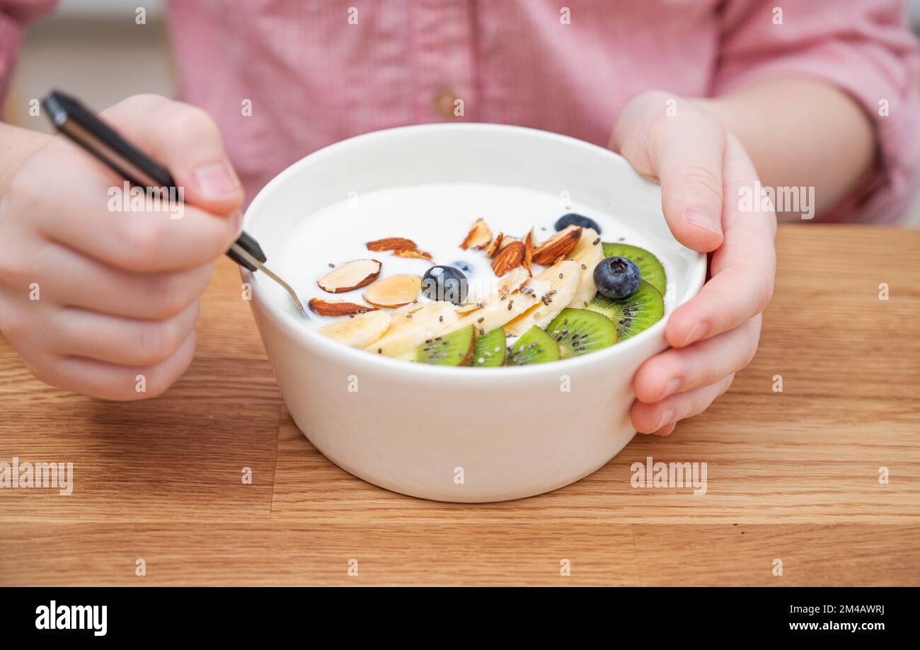 The hands of a girl in a pink shirt hold a bowl of yogurt, granola and fresh fruit on a wooden table in close-up. The concept of healthy eating. Stock Photo