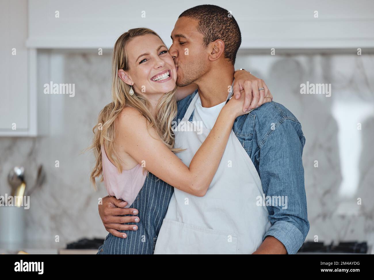 Love, interracial and couple kiss in kitchen, happiness and loving together for relationship. Romance, man and woman with embrace, kissing and Stock Photo