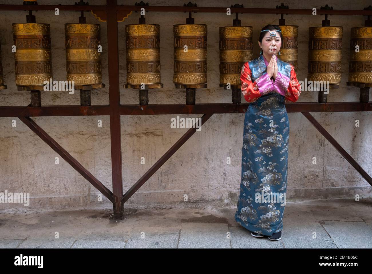 A Chinese tourist poses for a photo wearing traditional Tibetan clothing in front of prayer wheels. Lhasa. Tibet Autonomous Region. China. Stock Photo
