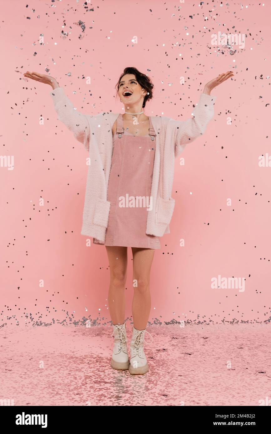 full length of joyful woman in fluffy cardigan catching confetti with raised hands on pink Stock Photo