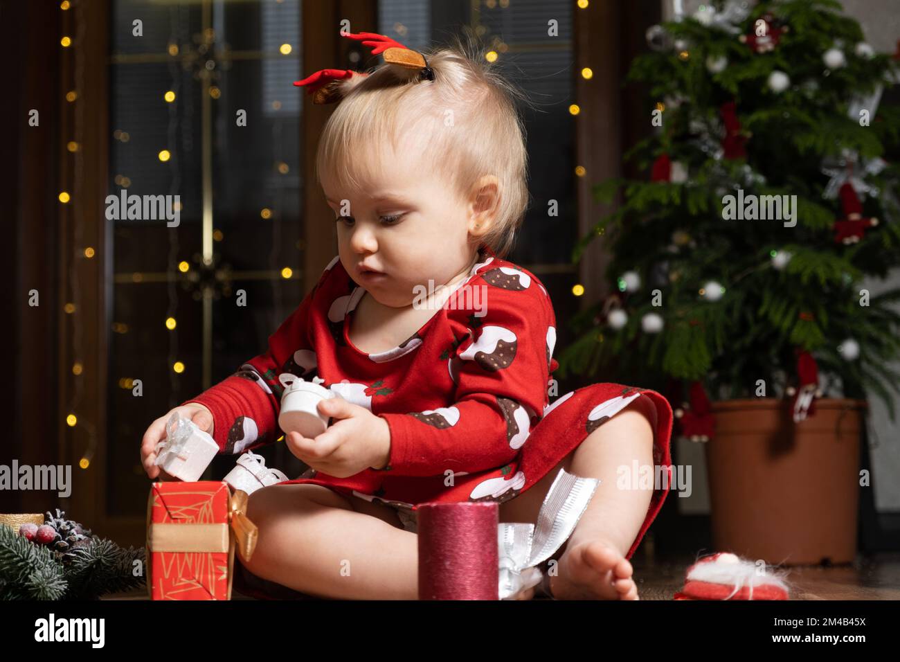 Beautiful little one-year-old girl with blonde hair playing Christmas gifts. Bokeh garlands. Children and new year's eve Stock Photo