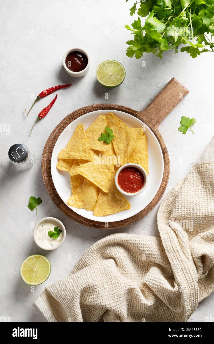 Corn chips nachos with salsa and cheese sauce, fresh herb, lime and chili pepper on white background. Concept Mexican food. Top view and copy space. Stock Photo