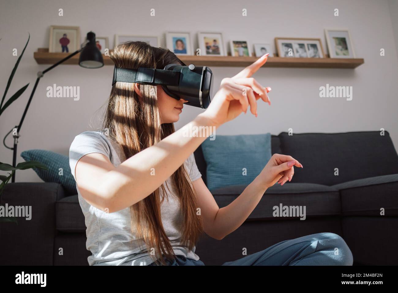 Young woman wearing virtual reality glasses, exploring the augmented world, interacting with the digital interface while sitting in the living room Stock Photo
