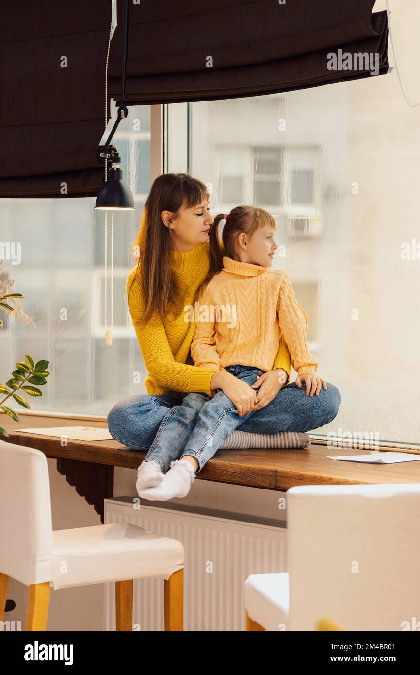 Mom and daughter hugging sitting near the window. Mom and child spend time together. Affectionate mother Stock Photo