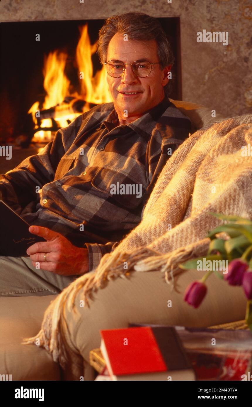 Older man sitting on the couch in front of a fireplace pausing to look up while reading a book Stock Photo