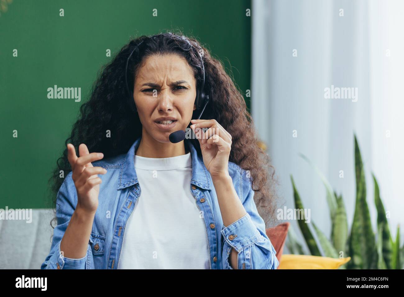 Portrait of angry woman with headset at home, Hispanic woman working remotely as consultant, looking displeased at camera video call online consultation.. Stock Photo