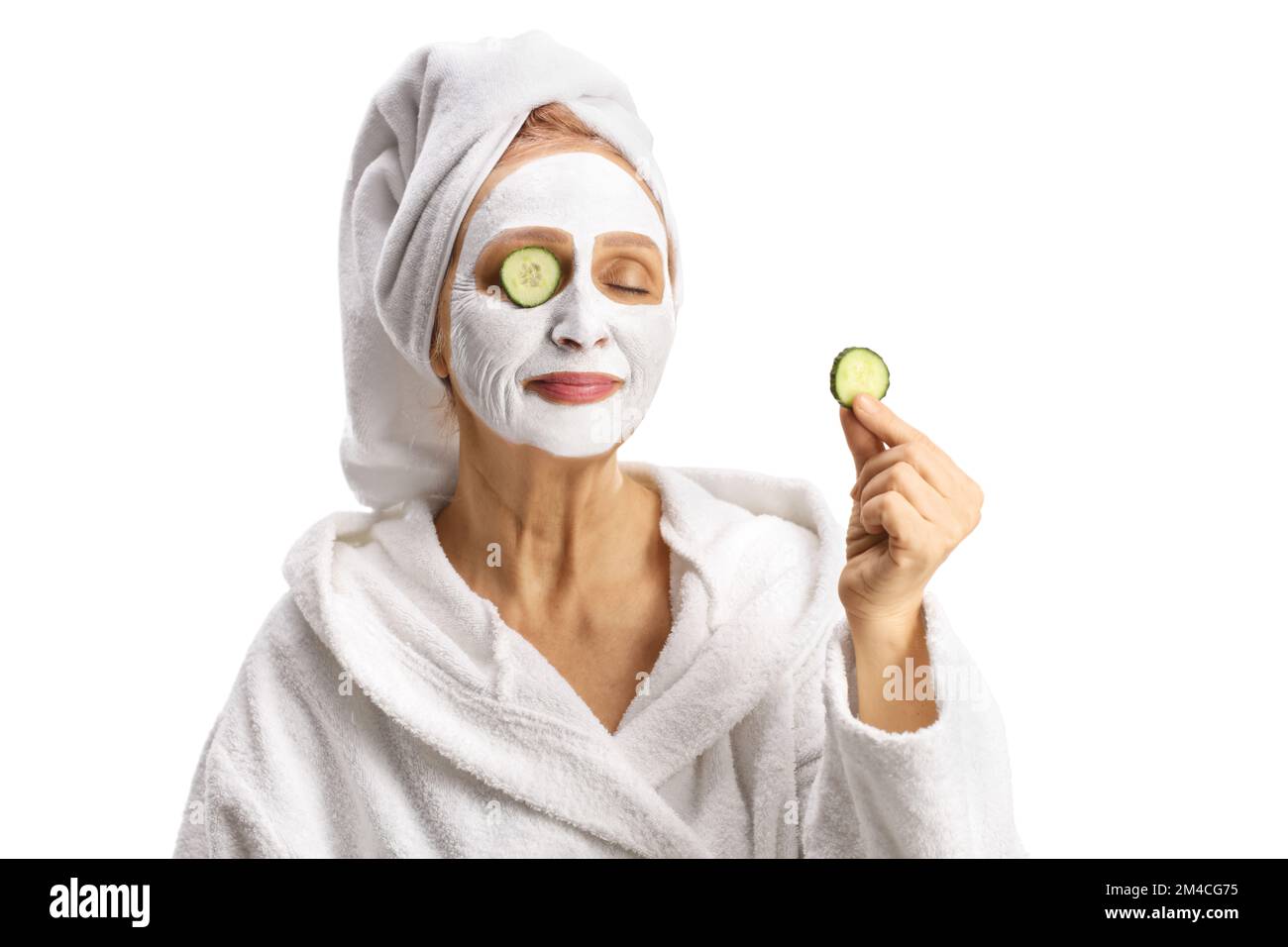 Woman with a towel on her head and a face mask holding a piece of cucumber isolated on white background Stock Photo